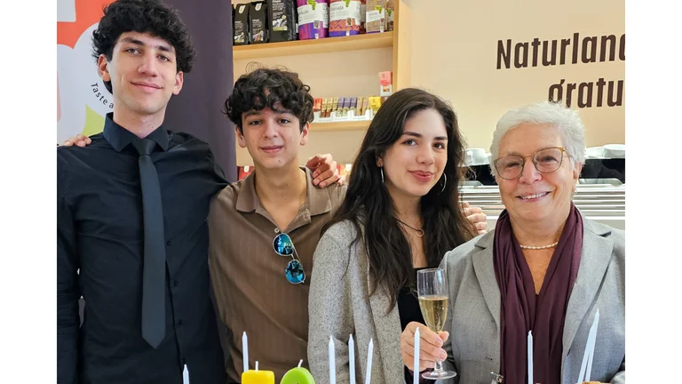 Zwei junge Männer, eine junge Frau und eine ältere Frau vor einem Messestand mit Kaffeepackungen Schokoriegeln lächeln in die Kamera. Die ältere Frau hält ein Glas Sekt in der Hand.