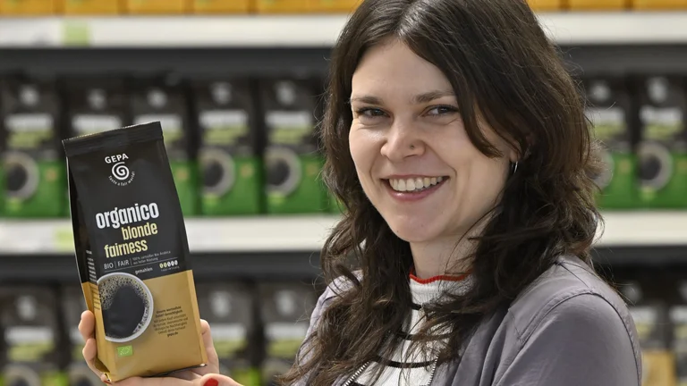 Sortimentsmanagerin Hanna Schmid mit Kaffeepackung "organico blonde fairness" in der Hand vor Regal mit weiteren Organico-Varianten.