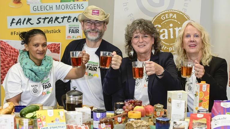 Drei Frauen und ein Mann sitzen an einem üppig gedeckten Frühstückstisch inkl. GEPA-Produkten. Sie prosten mit Tee in Richtung Kamera. Im Hintergrund sind Banner der GEPA zu sehen.