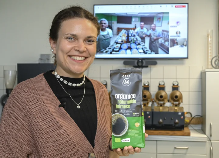 Eine Frau in einem Kaffeelabor lächelt und hält eine Packung GEPA-Kaffee "organico" in die Kamera. Hinter ihr auf einem Bildschirm sind per Videocall drei Männer zugeschaltet, die sich auch in einem Kaffeelabor befinden.