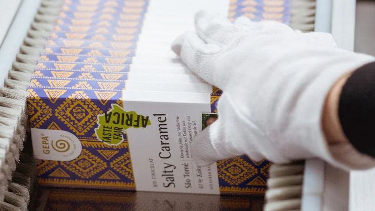 Hand mit weißem Handschuh entnimmt verpackte GEPA-Schokotafeln aus der Maschine.