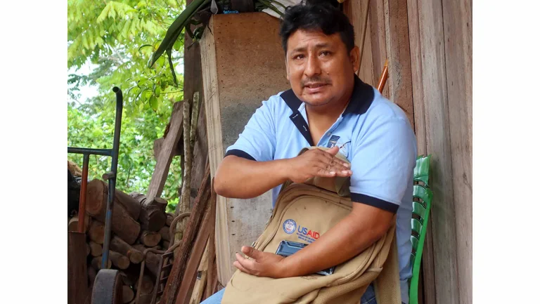Mann in hellblauem Polohemd sitz auf einem Plastikstuhl an einer Holzh&uuml;tte und hat einen Rucksack auf dem Scho&szlig;. Er schaut in die Kamera.