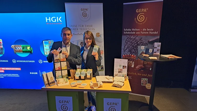 Mann und Frau an einem Messestand. Sie schauen in die Kamera und pr&auml;sentieren GEPA-Produkte wie Tee und Schokolade.