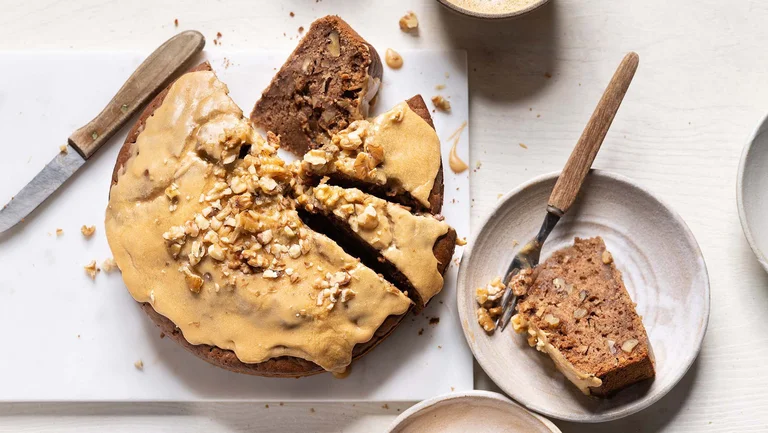Geschnittener Walnusskuchen mit beigem Topping auf einer Platte, daneben ein Stück Kuchen auf einem Teller mit Gabel