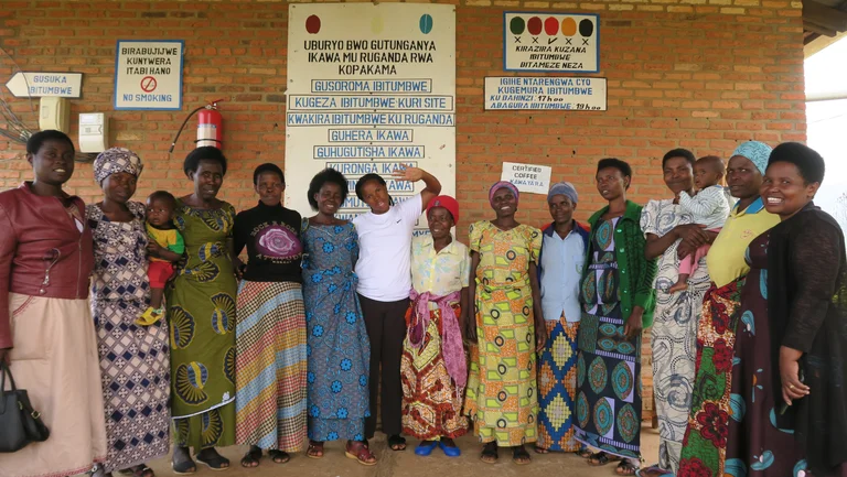 Eine Gruppe von 13 Frauen steht vor einer gemauerten Hauswand mit Schildern darauf und schaut in die Kamera. Zwei von ihnen tragen ein Kind auf dem Arm.