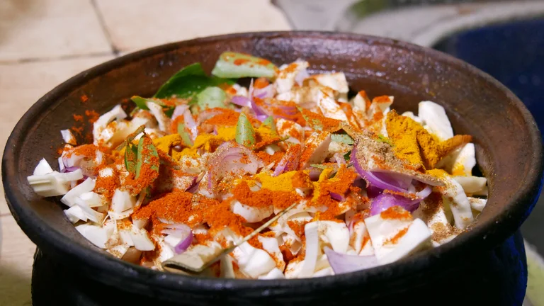 Jackfruit mit Zwiebeln und anderem Gem&uuml;se in einer dunklen Pfanne, darauf verteilt ein r&ouml;tliches Gew&uuml;rz-Pulver.
