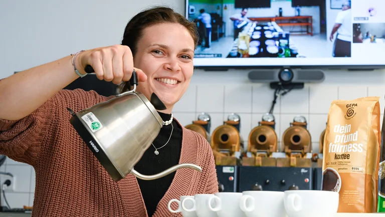 Junge Frau in einem Kaffeelabor hält eine silberne Kanne über weiße Tassen, als würde sie Kaffee aufgießen, und lächelt in die Kamera. Am Rand steht eine Packung GEPA-Kaffee "dein pfund fairness".