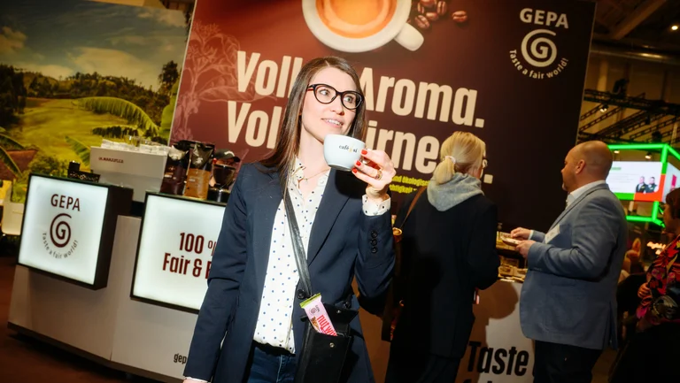Junge Frau mit Kaffeetasse in der Hand, aus ihrer Handtasche guckt ein GEPA-Nussriegel heraus, im Hintergrund der GEPA-Messestand