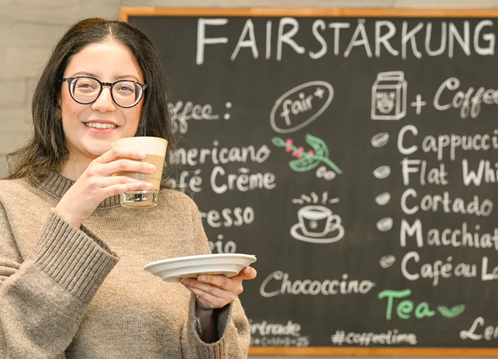 Junge Frau mit dunklen Haaren mit Kaffee vor einer Tafel mit dem Schriftzug "Fairstärkung"
