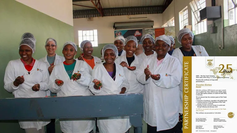 Elf Frauen in weißer Arbeitskleidung und mit Haarnetzen lächeln in die Kamera. Sie halten kleine rote Chilischoten in den Händen. Unten rechts in das Bild eingeklinkt ist ein Zertifikat über 25 Jahre Partnerschaft mit der GEPA.