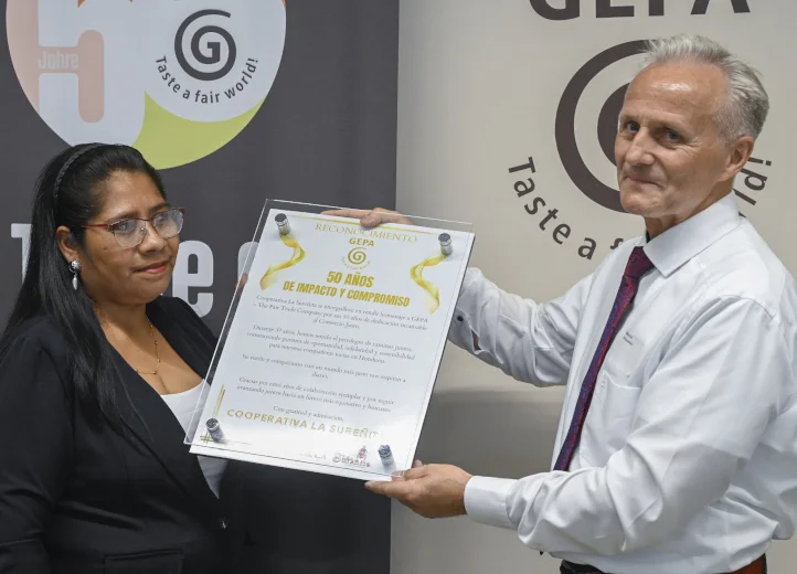 Frau und Mann stehen vor Bannern mit GEPA-Logo. Er zeigt eine Urkunde in die Kamera mit der Aufschrift „50 años de impacto y compromiso“