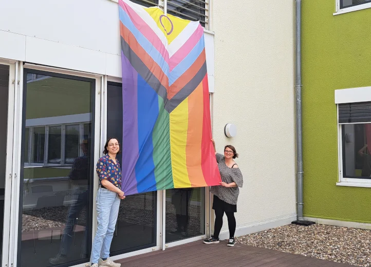 Eine große Pride-Regenbogenfahne hängt aus den Fenstern eines Büro des GEPA-Gebäudes. Zwei junge Frauen halten je eine Ecke der Fahne fest und lächeln in die Kamera.