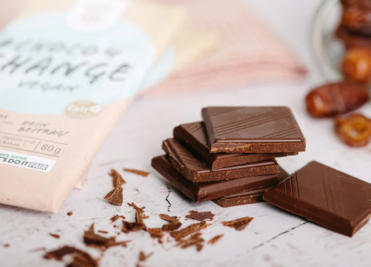 Ein Stapel Schokoladenstücke neben einer Tafel #Choco4Change Vegan und Schokosplittern