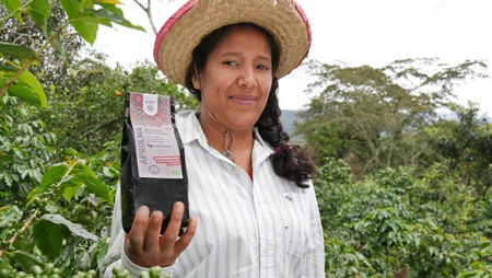 Frau h&auml;lt ein P&auml;ckchen GEPA-Kaffee "APROLMA" in die Kamera, in ihrem Feld stehend.