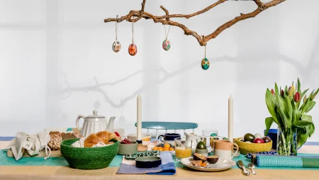 Bunt gedeckter Fr&uuml;hst&uuml;ckstisch u.a. mit Kaffeekanne, Kerzen, bunten Eier und einer Vase mit Tulpen. Dar&uuml;ber h&auml;ngt ein Ast, von dem bunt bemalte Ostereier herunterh&auml;ngen.