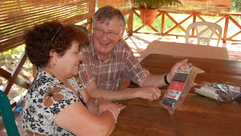 Mann und Frau sitzen an einem Tisch auf einer Art Veranda. Er zeigt ihr eine alte Packung Kaffee und lacht sie an.
