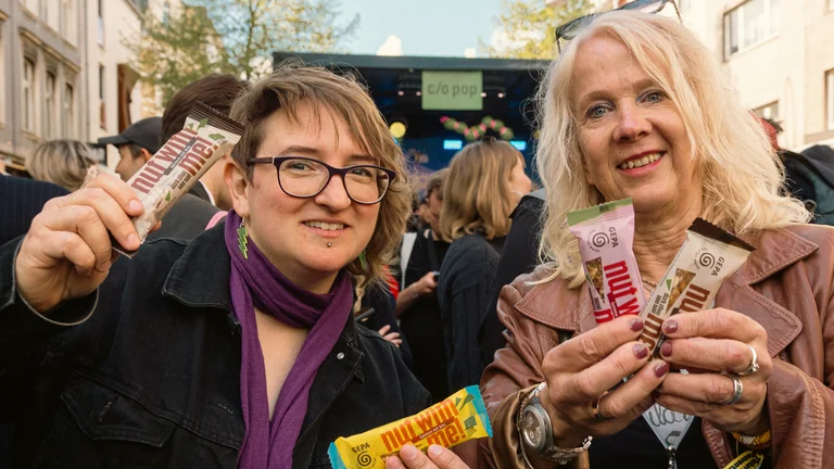 Barbara Schimmelpfennig und Anne Bien halten GEPA-Nussriegel in die Kamera, hinter ihnen eine Open-Air-B&uuml;hne und Konzertg&auml;ste. 