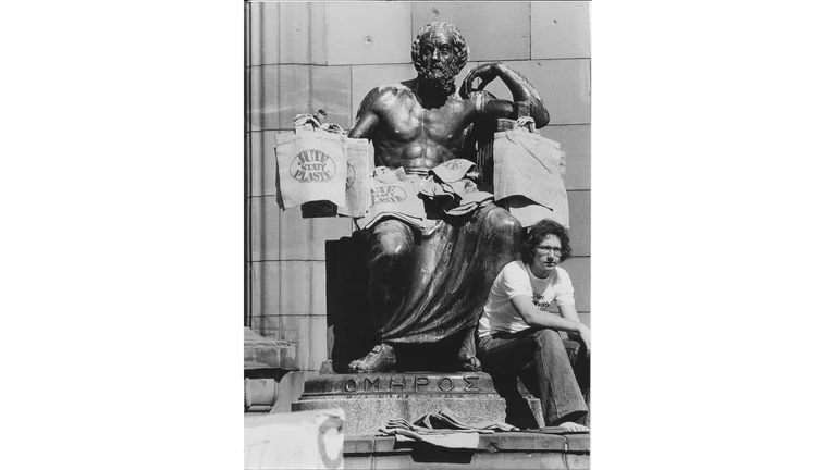 Schwarz-Wei&szlig;-Aufnahme: Auf einer sitzenden Statue eines Mannes liegen Jutetaschen. Am Sockel sitzt ein junger Mann mit Locken und Brille.