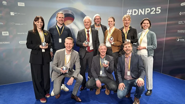 Gruppenfoto mit 10 Personen auf einem blauen Teppich. Jede Person hat eine Preisstatuette in der Hand, eine silberfarbene Kugel. 