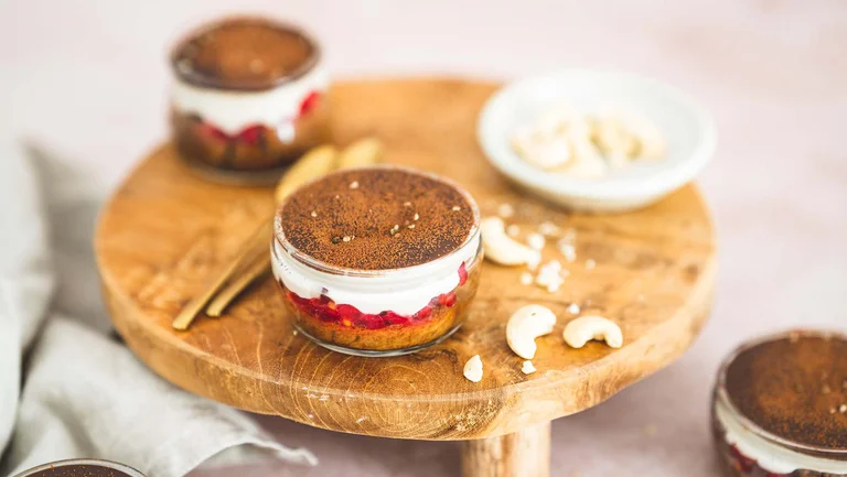 Ein Gläschen buntes Tiramisu auf einer Holzplatte mit Löffel und Cashews daneben