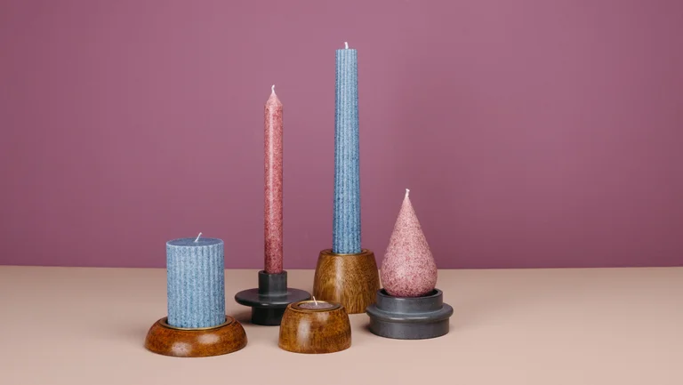 Fünf Kerzen in verschiedenen Farben und Formen stehen in Kerzenständern vor einer violetten Wand.