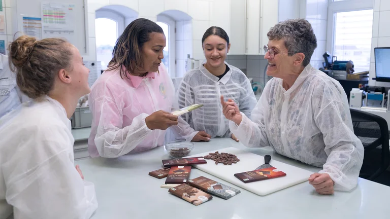 Vier Frauen stehen um einen Tisch und unterhalten sich lebhaft. Eine h&auml;lt eine Tafel Schokolade in der Hand. Auf dem Tisch liegen weitere Tafeln und Kakaobohnen. Alle tragen d&uuml;nne Kittel &uuml;ber ihrer Kleidung.