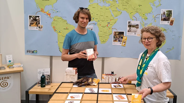 Junger Mann und &auml;ltere Frau an einem Messestand l&auml;cheln in die Kamera. Hinter ihnen ist eine Weltkarte aufgeh&auml;ngt. Vor ihnen liegt eine Art Schachbrett aus Holz. Beiden halten GEPA-Produkte in den H&auml;nden.