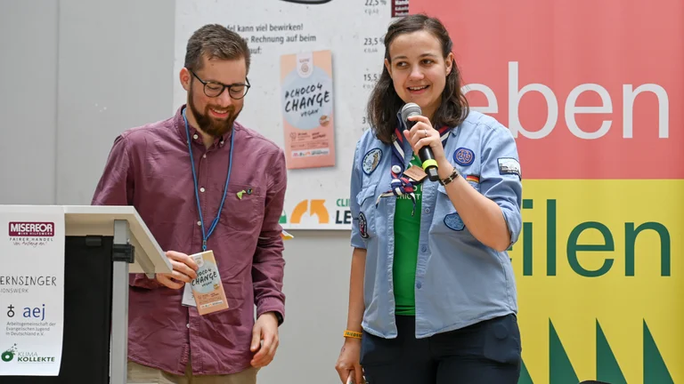 Junger Mann und junge Frau in Pfadfinder-Klamotten auf einer B&uuml;hne. Er h&auml;lt eine Schokolade "#Choco4Change Vegan" in der Hand, sie spricht in ein Mikrofon. Auf einem Plakat hinter ihnen ist die gleiche Schokolade zu sehen.