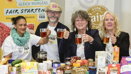 Drei Frauen und ein Mann sitzen an einem &uuml;ppig gedeckten Fr&uuml;hst&uuml;ckstisch inkl. GEPA-Produkten. Sie prosten mit Tee in Richtung Kamera. Im Hintergrund sind Banner der GEPA zu sehen.
