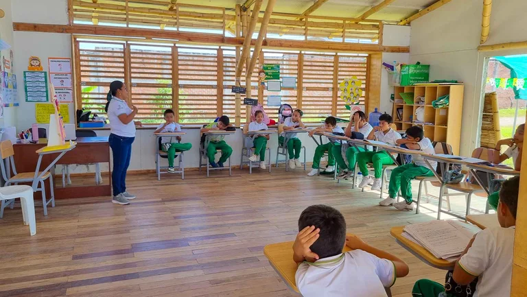 In einem aus Bambus gebauten Klassenzimmer gibt eine Lehrerin Unterricht. In U-Form sitzen um sie herum ca. 15 Schüler*innen an ihren Tischen.
