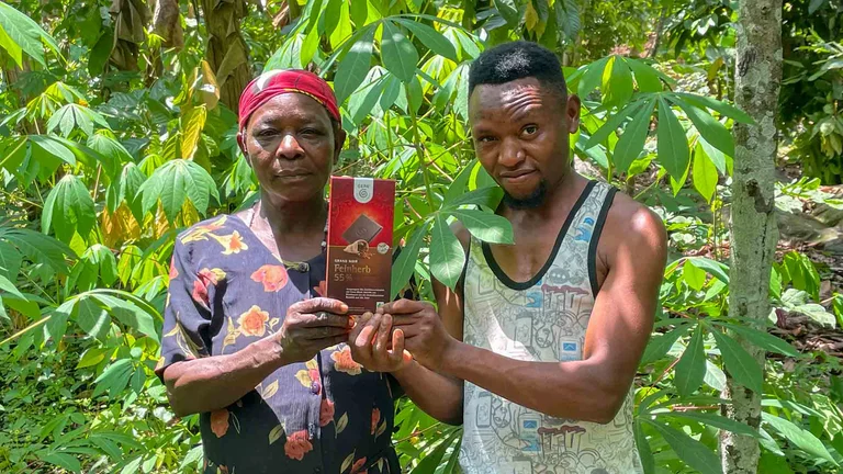 Zwei Personen umringt von gr&uuml;nen Bl&auml;ttern halten eine Tafel GEPA-Schokolade Feinherb 55 % in die Kamera.