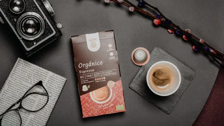 Auf einem Tisch liegt eine Packung GEPA-Kaffeekapseln "Org&aacute;nico Espresso". Daneben eine Brille auf einer Zeitung, eine Kamera und eine Tasse Kaffee auf einem Schieferuntersetzer.