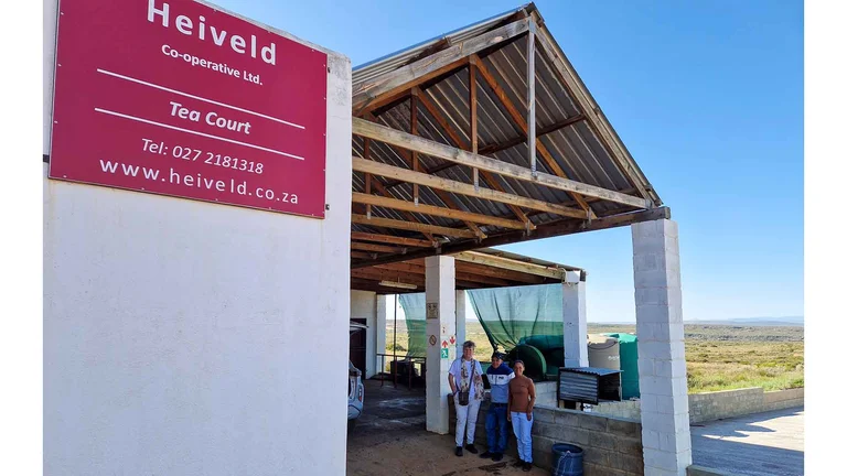 Gebäude vor einem weiten Feld. In einer Art offenem Carport stehen drei Personen. An einer Hauswand im Vordergrund hängt ein rotes Schild, auf dem steht: "Heiveld Co-Operative Ltd. Tea Court"