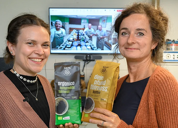 Zwei Frauen in einem Kaffeelabor lächeln und halten je eine Packung GEPA-Kaffee in die Kamera. Hinter ihnen auf einem Bildschirm sind per Videocall drei Männer zugeschaltet, die sich auch in einem Kaffeelabor befinden.