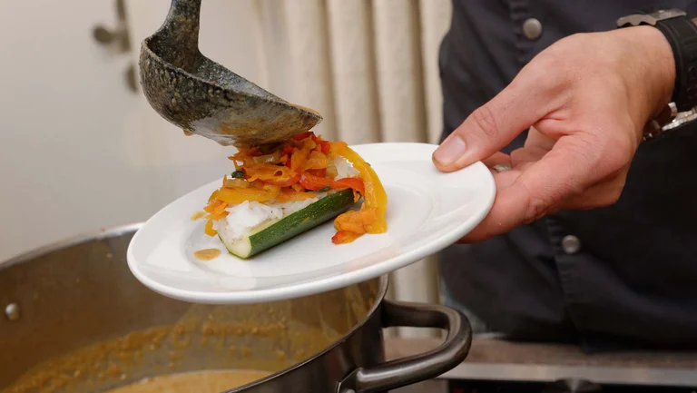 Eine Hand hält einen Teller mit einer Zucchini darauf. Aus einer Kelle kommt Gemüse darauf