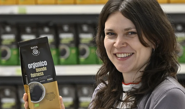 Sortimentsmanagerin Hanna Schmid mit Kaffeepackung "organico blonde fairness" in der Hand vor Regal mit weiteren Organico-Varianten.