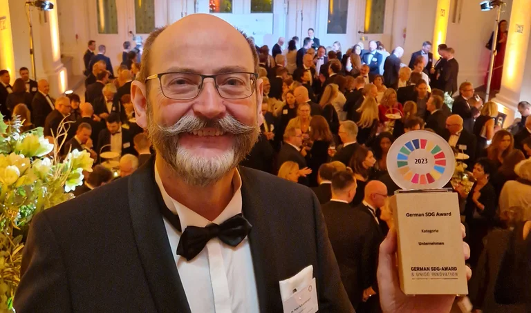 GEPA-Geschäftsführer P. Schaumberger hält den SDG-Award aus Holz in der Hand, hinter ihm der rote Teppich und ein Blick auf die Feier.