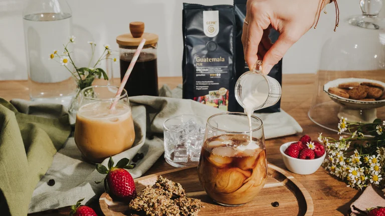 Auf einem Tisch sind ein Tuch, Erdbeeren, Kekse unter einer Gllasglocke, Blumen, Himbeeren in einer Schale und mehr angerichtet. Eine Hand gießt Milch aus einem kleinen Messbecher in ein Glas mit Kaffee und Eiswürfeln. Im Hintergrund sind zwei Packungen GEPA-Kaffee zu erkennen.