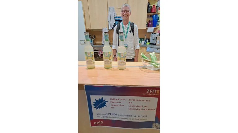 Mann steht hinter einer Theke und l&auml;chelt in die Kamera. Auf dem Tresen stehen drei Flaschen GEPA-Limonade und eine Sch&uuml;ssel mit Sesamriegeln darin.