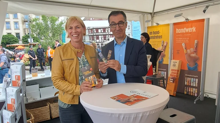 Cem &Ouml;zdemir und eine Frau stehen an einem Stehtisch an einem Messestand der GEPA. Sie halten GEPA-Schokoriegel bzw. einen GEPA-Flyer in H&auml;nden.