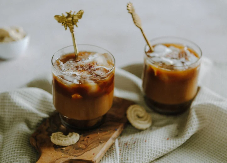 Zwei Gläser gefüllt mit Eiskaffee und einem Löffel darin auf einem Tuch, dekoriert mit Keksen