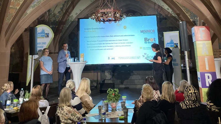 Blick aus dem Publikum auf eine B&uuml;hne, die in einer Kirche steht. Auf der B&uuml;hne stehen ein Mann und drei Frauen neben einer Leinwand. Darauf eine Folie mit der &Uuml;berschrift "unsere Gesellschafter und ihre Ziele".