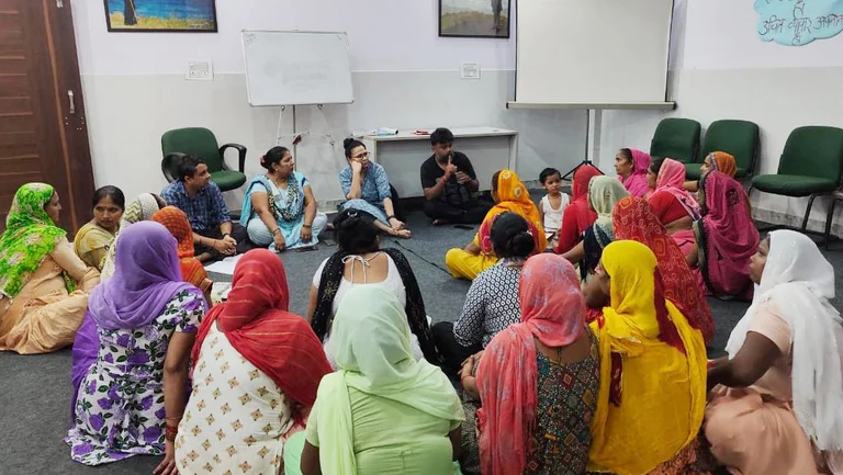 Gruppe von ca. 20 Menschen, meist Frauen in Saris, sitzt in einem Seminarraum im Kreis auf dem Boden und diskutiert.