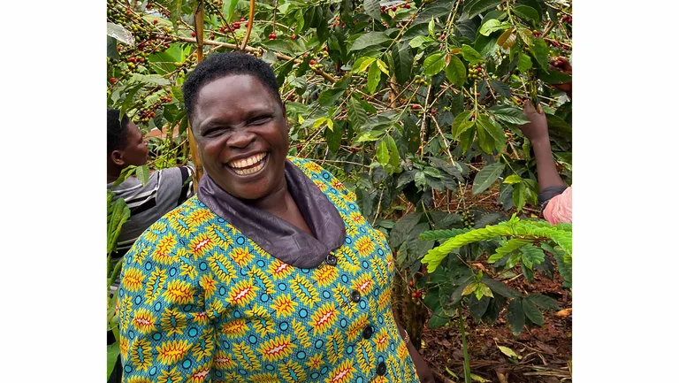 Frau in einem bunten Oberteil steht in einer Kaffeepflanzung und lacht in die Kamera.