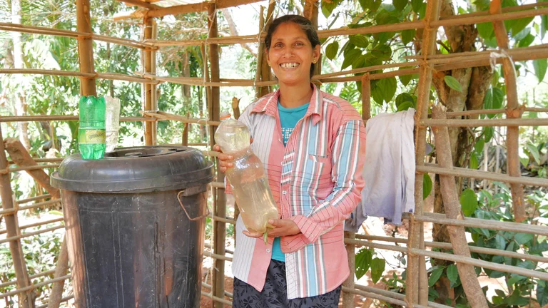 Sumithra steht mit einer gefüllten Flasche neben einer schwarzen runden Tonne, an der wiederum mehrere Flaschen mit dem Hals nach unten eingesteckt sind.