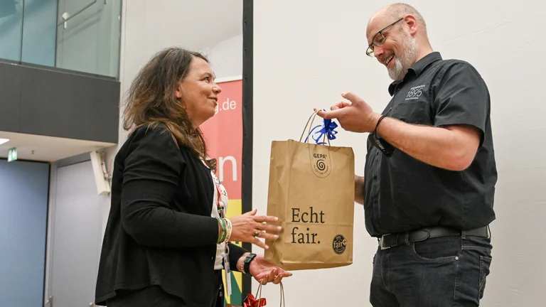 Frau &uuml;berreicht Mann eine braune Geschenkt&uuml;te aus Papier mit GEPA-Logo und der Aufschrift "Echt fair."