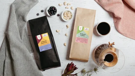 Man sieht von oben, wie zwei Packungen Kaffee (eine schwarz, eine hellbraun) auf einem Tisch liegen. Daneben liegen eine Schale mit N&uuml;ssen, eine mit Beeren sowie eine Tasse und eine Chemex mit Kaffee gef&uuml;llt.