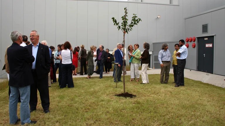 Gruppe von ca. 25 Menschen steht auf einer Gr&uuml;nfl&auml;che vor einem grauen Geb&auml;ude. Auf der Gr&uuml;nfl&auml;che steht ein frisch gepflanzter junger Kirschbaum.  