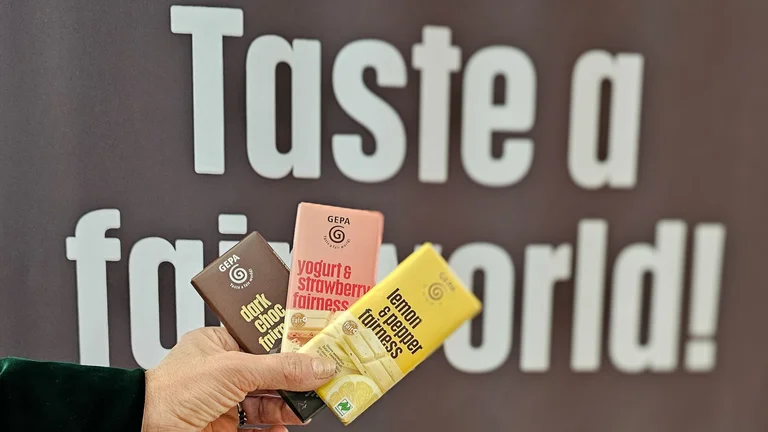 Eine Hand hält drei verschiedene GEPA-Schokoladen. Dahinter ist groß der Schriftzug "Taste a fair world!" zu erkennen.