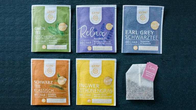 Sechs Sorten GEPA-Teebeutel, angeordnet in zwei Reihen in ihren Kuverts, mit Ausnahme unten rechts: Dieser Beutel ist ausgepackt.